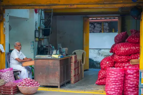A small shop owner writing in a notebook behind a counter stacked with goods